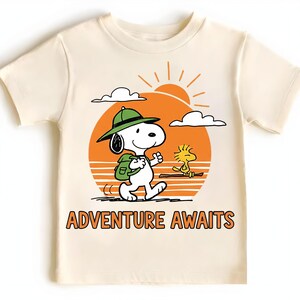 May include: Cream-colored t-shirt featuring a cartoon Snoopy character wearing a green hat and backpack, walking towards the right. A yellow Woodstock character is also present. The shirt has an orange sun graphic and the words "ADVENTURE AWAITS" in orange.