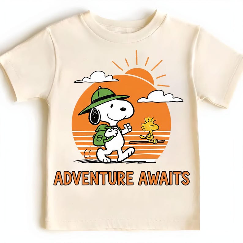 May include: Cream-colored t-shirt featuring a cartoon Snoopy character wearing a green hat and backpack, walking towards the right. A yellow Woodstock character is also present. The shirt has an orange sun graphic and the words "ADVENTURE AWAITS" in orange.