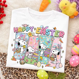 May include: White t-shirt with a colorful "Happy Easter" graphic featuring Snoopy characters. The design includes Easter eggs, carrots, flowers, and Snoopy wearing bunny ears. A festive and cheerful design.