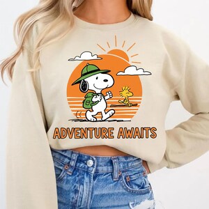 May include: Beige sweatshirt featuring a graphic of Snoopy with a backpack and Woodstock. The design includes an orange sun, clouds, and the text "ADVENTURE AWAITS". The sweatshirt is a neutral color.