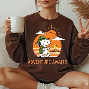 May include: Brown sweatshirt featuring Snoopy and Woodstock walking towards a sunset with the words "Adventure Awaits." Snoopy is wearing a green hat and backpack. The design is printed in orange, white, and black.