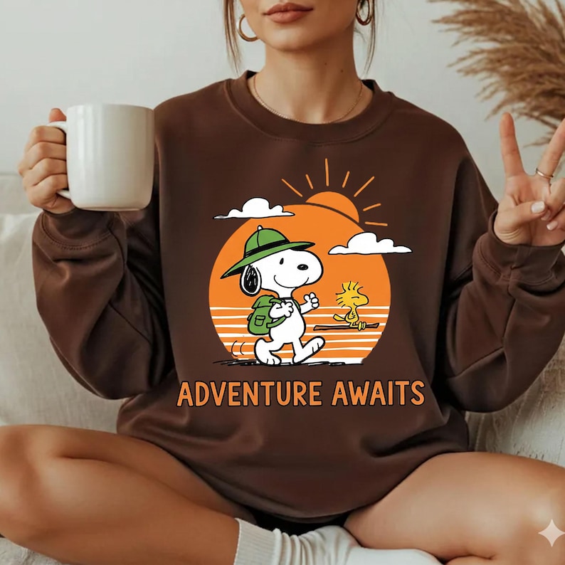 May include: Brown sweatshirt featuring Snoopy and Woodstock walking towards a sunset with the words "Adventure Awaits." Snoopy is wearing a green hat and backpack. The design is printed in orange, white, and black.