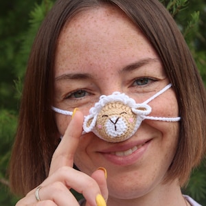 May include: A crocheted sheep nose accessory, with a tan face, white wool, and pink accents. The sheep is attached to a white string that goes around the ears. The accessory is worn on the nose.