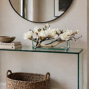 May include: A modern entryway featuring a round black-framed mirror, a glass console table, and a woven basket. The table displays a bowl, books, and a floral arrangement. A wall sconce with a lit candle adds warmth.