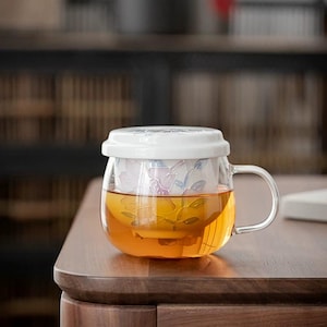 May include: A clear glass teacup with a white lid and a floral design. The teacup has a handle and is filled with amber-colored tea.