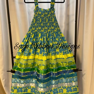 May include: A yellow and blue floral print dress with ribbon accents. The dress has a smocked bodice and a full skirt with horizontal stripes of yellow, green, blue, and white ribbons. The text "Sage Ribbon Designs" is visible.