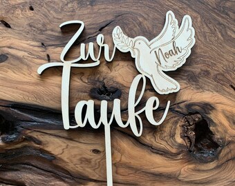 Może przedstawiać: Drewniany topper na tort z napisem "Zur Taufe" i gołębiem z gałązką oliwną w dziobie. Gołąb ma napisane imię "Noah".