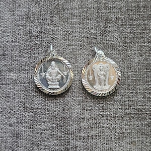 Puede incluir: Dos colgantes plateados con figuras religiosas. Cada colgante redondo tiene un borde texturizado. Uno muestra una figura sentada, el otro de pie. Ambos tienen un pequeño lazo en la parte superior para una cadena.