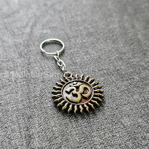 Puede incluir: Un llavero plateado con un dije redondo en forma de sol que presenta el símbolo Om en marrón y dorado. El dije tiene un diseño texturizado en forma de sol.