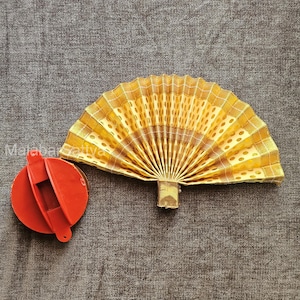 May include: A decorative, open fan with a gold and yellow patterned design. The fan is made of pleated fabric and has a wooden handle. A red, circular object with a hinged lid is next to the fan.