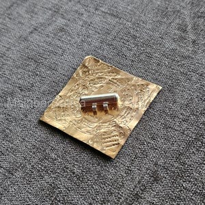 May include: A gold-colored, square-shaped item with intricate embossed patterns and a silver cylindrical object in the center. The item is resting on a gray, textured surface.