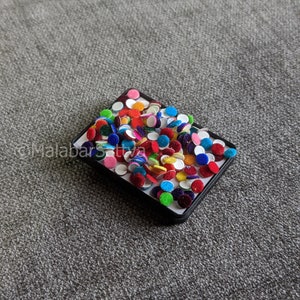 Puede incluir: Un pequeño contenedor negro lleno de adornos redondos de colores. Los adornos son de varios colores, incluyendo rojo, azul, verde, amarillo y blanco.