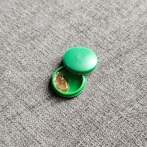 May include: A small, open green container with a matching lid. Inside the container is a small amount of a light brown, semi-transparent substance. The container is on a grey, textured surface.