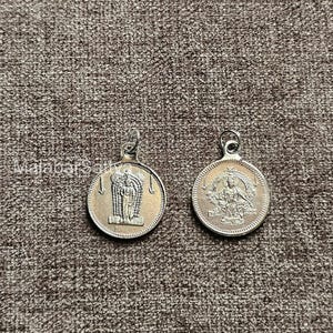 Puede incluir: Dos colgantes plateados con figuras religiosas. Cada colgante circular presenta una imagen detallada de una deidad. Los colgantes tienen un lazo en la parte superior para una cadena o cordón.