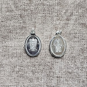 Puede incluir: Dos colgantes ovalados plateados con diseños detallados. Uno presenta una imagen arquitectónica, el otro una figura sentada. Ambos tienen un pequeño lazo para una cadena. Fondo texturizado y neutro.