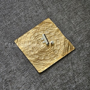 Puede incluir: Un cuadrado de color dorado con patrones y escritura intrincados, un tubo plateado y una pequeña bola plateada.