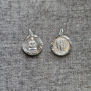 Puede incluir: Dos colgantes plateados con figuras religiosas. Uno presenta un Buda sentado, el otro una figura de pie. Ambos son circulares con un borde texturizado y un lazo para una cadena. Se muestran sobre una superficie gris texturizada.