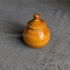 Puede incluir: Un pequeño recipiente redondo de madera con tapa. El recipiente es de color marrón claro y tiene una franja blanca alrededor del medio. La tapa es de color marrón oscuro y tiene una parte superior redondeada.