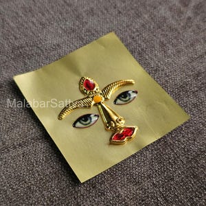 Puede incluir: Una pegatina de color dorado con una cara estilizada que presenta piedras preciosas rojas y azules para los ojos y los labios. La pegatina está sobre un fondo amarillo.