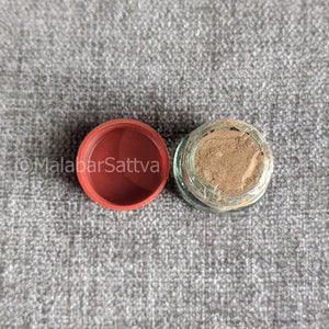 Può includere: Un piccolo barattolo di vetro con un coperchio di plastica rosso. Il barattolo è pieno di una polvere marrone chiaro. Il barattolo è su una superficie grigia testurizzata.