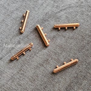 May include: Five small, cylindrical, copper-colored metal beads with three small, protruding pegs on each side. The beads are scattered on a textured, gray surface. The beads have a shiny, metallic finish.