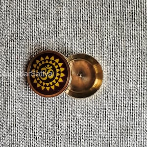Puede incluir: Un pequeño recipiente redondo de latón con tapa. La tapa tiene un diseño amarillo y marrón con el símbolo "Om" en el centro.