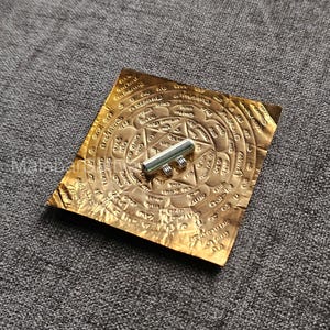 May include: A silver amulet with a cylindrical shape and a decorative design. The amulet is resting on a gold-colored square piece of paper with intricate patterns and writing in a foreign language.