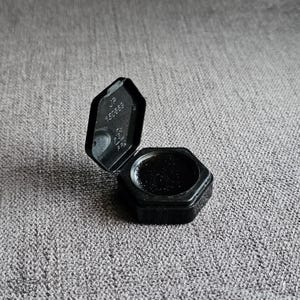 以下が含まれることがあります： 開いたヒンジ付きの蓋が付いた小さな黒い六角形の容器。蓋の内側には、エンボス加工されたテキスト「JP 150889」とリサイクルマークがあります。容器の底部には、六角形のフレーム内に暗い質感のある物質が入っています。