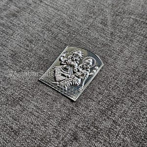 May include: A silver pendant featuring a detailed depiction of a Hindu deity couple, likely Shiva and Parvati, in a traditional pose. The pendant is rectangular with rounded edges and has a textured surface.