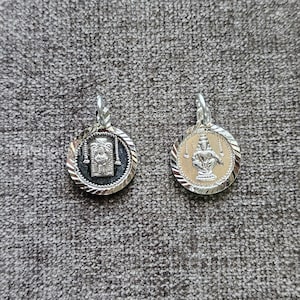 Puede incluir: Dos colgantes plateados con diseños detallados. Uno presenta una imagen dentro de un marco rectangular, el otro una figura sentada. Ambos son circulares con un borde texturizado y un lazo para una cadena. Para collares.