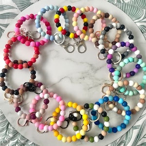 May include: A selection of beaded keyrings in a circular arrangement. The keyrings display a variety of colours, such as pink, blue, yellow, and green. Each keyring has a metal clasp for attaching keys. The beads are made of a smooth, rounded material.