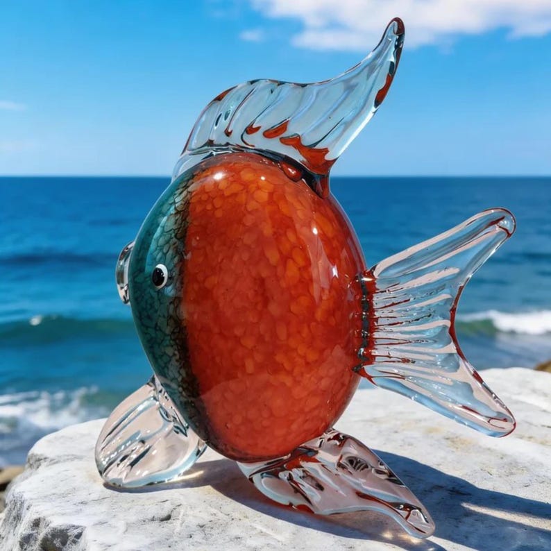 May include: A decorative glass fish sculpture with a vibrant orange body and clear fins. The fish has a textured surface and is displayed on a rock with the ocean and blue sky in the background. The sculpture is approximately 6 inches long.