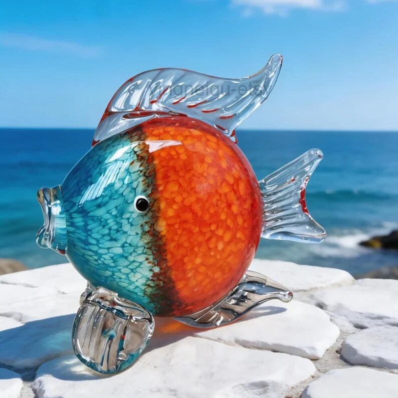 May include: A colorful glass fish sculpture with a vibrant design. The fish features a split color scheme, with one side in turquoise and the other in orange. Clear glass fins and tail complete the design. The sculpture is displayed on a white stone surface.