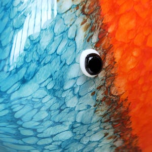 May include: Close-up of a colorful, abstract design featuring a white and black eye. The design has a textured appearance with shades of blue and orange. The eye is a prominent detail, adding a focal point to the artwork.