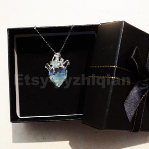 Puede incluir: Un collar de plata con un colgante de corazón de vidrio. El corazón es transparente con un degradado azul y verde. El collar está en una caja de regalo negra con una cinta dorada.
