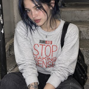 May include: A light gray long-sleeve crewneck sweatshirt featuring the text TALKING HEADS STOP MAKING SENSE in red and black lettering on the chest. The garment has ribbed cuffs and a ribbed neckline, paired with black pants and a black bag.