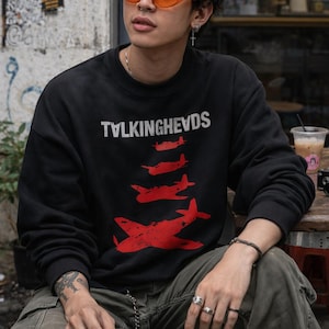 May include: Black sweatshirt with the text "TALKINGHEADS" and a red airplane graphic. The person is wearing orange sunglasses, cargo trousers, and rings. The person is sitting at a wooden table.