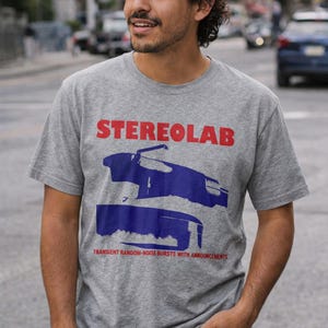 Può includere: Una maglietta grigia con la scritta "STEREOLAB" in rosso sopra una grafica blu. Sotto, il testo recita "TRANSIENT RANDOM-NOISE BURSTS WITH ANNOUNCEMENTS". La maglietta è indossata da una persona.