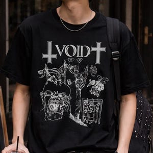 Puede incluir: Camiseta negra con un diseño gráfico blanco y la palabra "VOID" encima de una cruz. La camiseta presenta una ilustración detallada de figuras y símbolos. La persona lleva gafas de sol negras, una cadena plateada y un reloj.