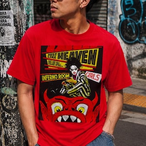 Può includere: Una t-shirt rossa a maniche corte con una stampa grafica che riporta il testo "JUST LIKE HEAVEN", "INFERNO ROOM" e "GIRLS". Il design include una figura stilizzata in abito a righe e un grande volto di diavolo rosso con occhi gialli.