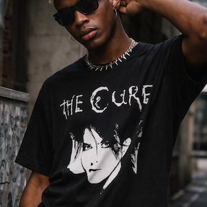 May include: A black short-sleeved t-shirt featuring a white graphic print of a face and the text THE CURE. The shirt has a crew neckline and is made of a solid black fabric. The graphic is centered on the chest, displaying a stylized portrait.