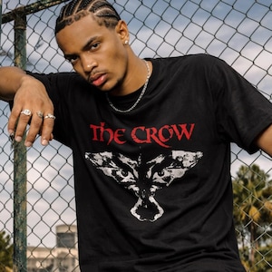 May include: A black short-sleeve t-shirt featuring the text THE CROW in red lettering above a white graphic illustration of a bird with spread wings. The shirt has a crew neckline and a standard fit, constructed from a solid black fabric material.