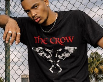 Puede incluir: Una camiseta negra de manga corta con el texto THE CROW en letras rojas sobre una ilustración gráfica blanca de un pájaro con las alas extendidas. La camiseta tiene cuello redondo y un corte estándar, confeccionada en tela negra lisa.
