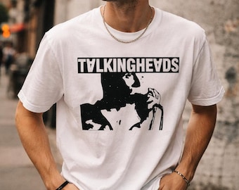 May include: White t-shirt with the band name "TALKINGHEADS" in black block letters above a black and white graphic. The shirt is worn with blue jeans and sunglasses.