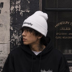 Può includere: Un berretto in maglia bianco con la parola philosophy ricamata in nero sul davanti, abbinato a una felpa con cappuccio nera caratterizzata da un design grafico bianco in un carattere stilizzato e frastagliato sul petto a sinistra.