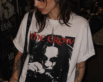 May include: A white short-sleeved t-shirt featuring a black and white graphic print of the character from the film The Crow, with the title text The Crow printed in bold red lettering across the top of the rectangular graphic design.