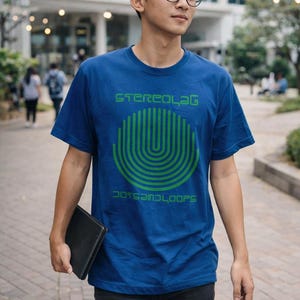 May include: A blue t-shirt with the text "STEREO LAG" and "DOTS AND LOOPS" in green. The shirt features a green, curved graphic design. The person in the image is wearing black pants and glasses.