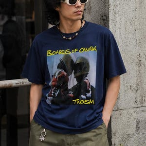 May include: Navy blue t-shirt with a graphic featuring the text "BOARDS OF CANADA" in yellow and an image of two figures. The shirt is paired with olive green trousers. The person is wearing sunglasses and a necklace.