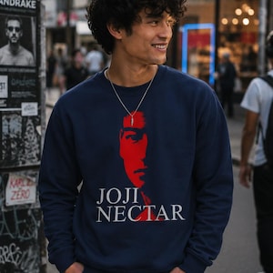 May include: A navy blue crewneck sweatshirt featuring a red graphic of a face and the white text JOJI NECTAR printed on the front. The garment has long sleeves and a ribbed neckline, cuffs, and hem, providing a standard fit for casual wear.