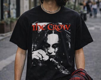 May include: Black t-shirt featuring the movie title "the Crow" in red lettering above a black and white image of a person. The shirt is worn with black jeans, a black cap, and round sunglasses. A red and black plaid shirt is draped over the arm.